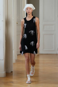 Model walking a fashion show in a black sleeveless dress with white fringe patches, white head wrap, and white crocheted sandals in an elegant, paneled-room setting.
