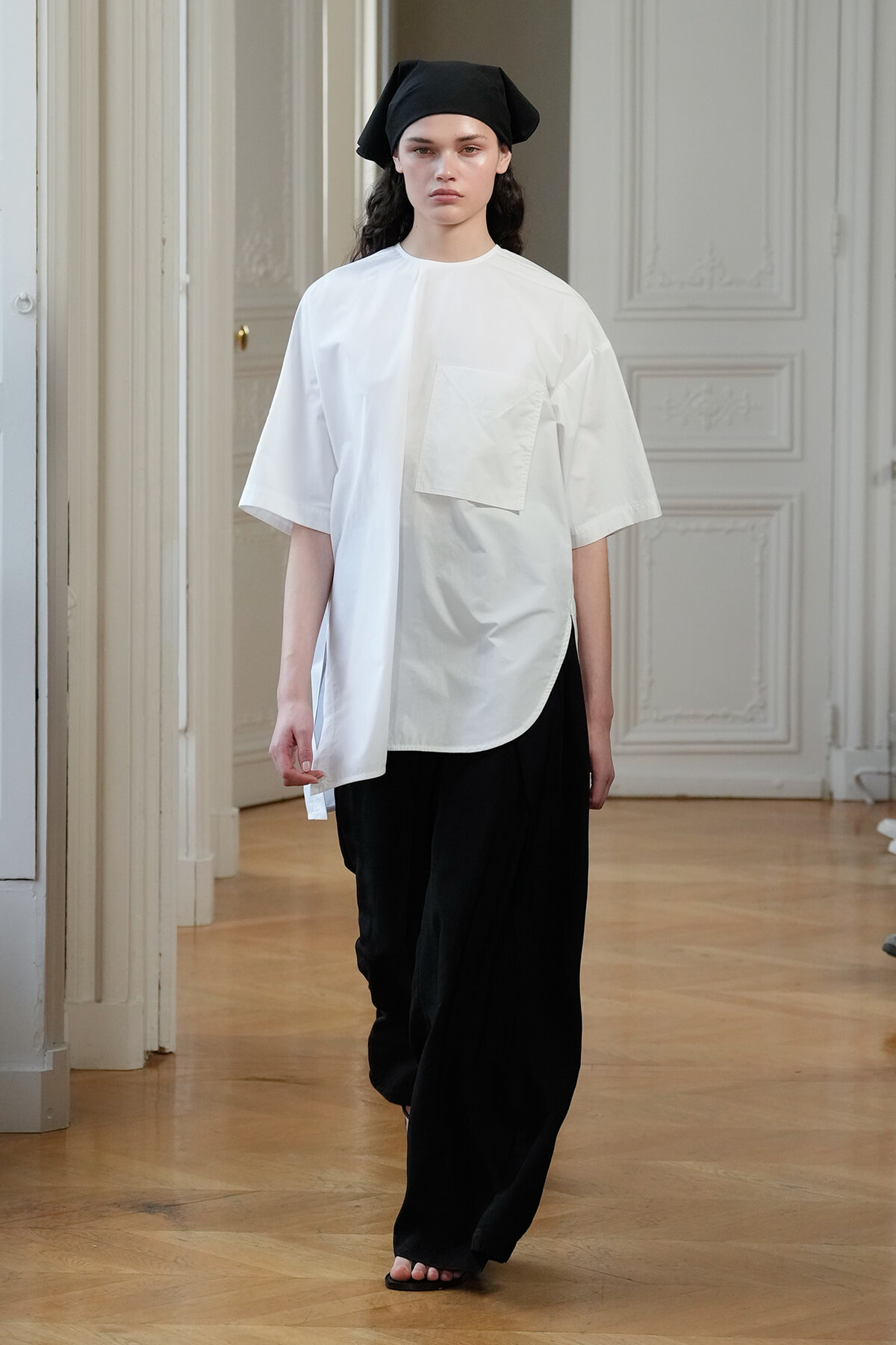 Model walking in a white oversized top with a large front pocket paired with black wide-leg pants and a black headscarf in a bright room.