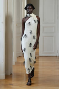 Model wearing a white knit, one-shoulder gown with black feather embellishments, paired with a black headscarf and sandals on an indoor runway.