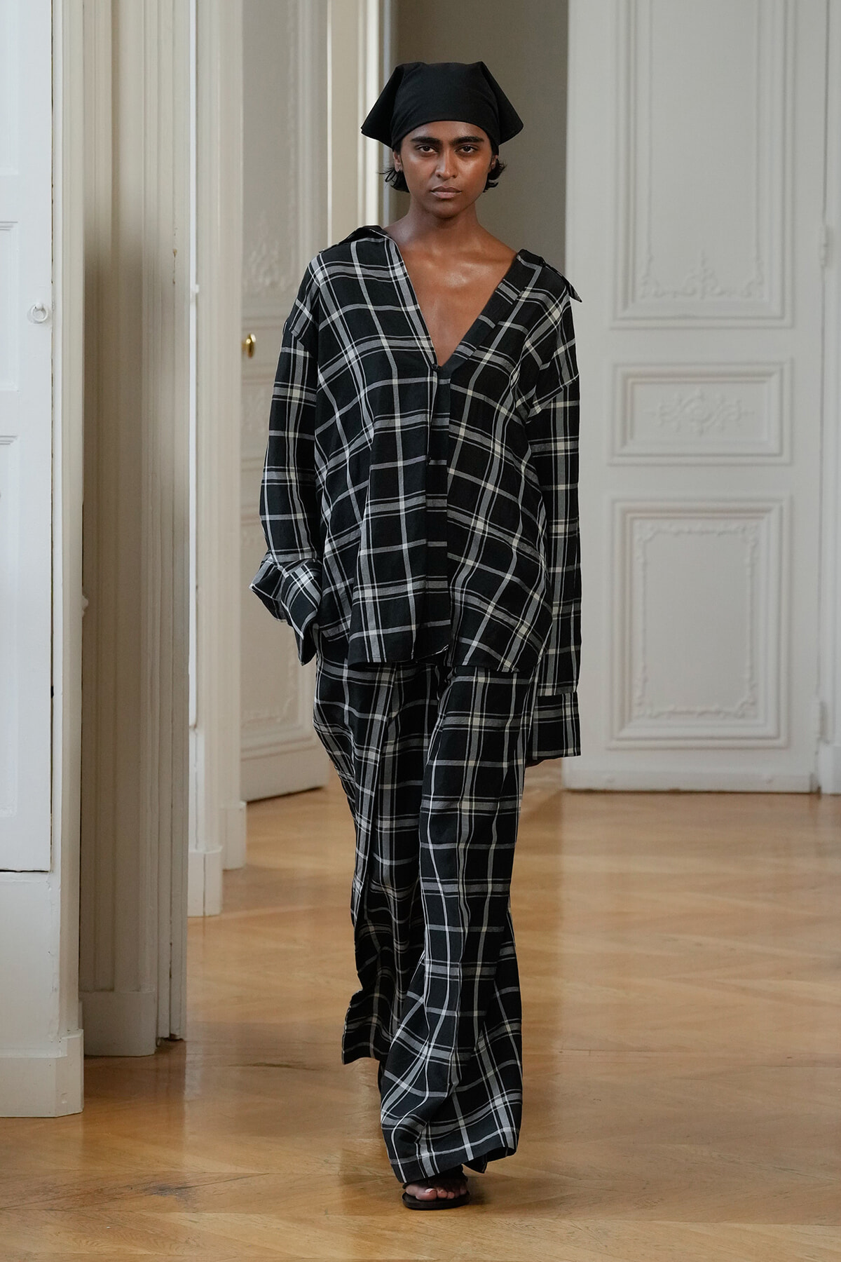 Model walking down a bright hallway in a black-and-white plaid oversized shirt and matching wide-leg pants, wearing a black head wrap.