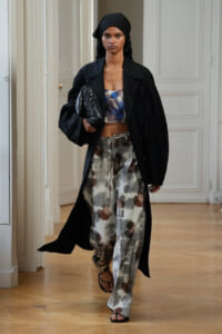 Model walks in an oversized black coat over a blue floral crop top and loose gray floral trousers, carrying a black clutch vessel.