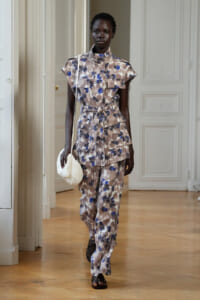 Model walking in a beige-blue floral-print jumpsuit with a waist belt, carrying a white bag in a bright, ornate room.