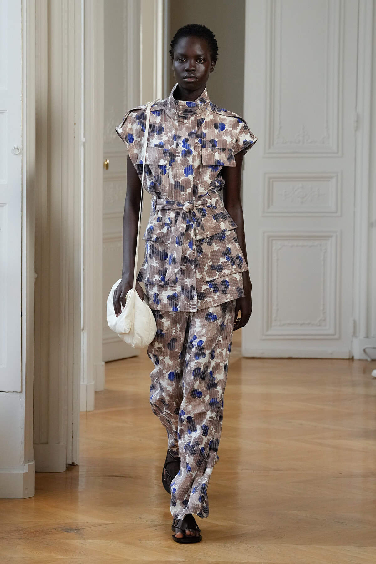 Model walking in a beige-blue floral-print jumpsuit with a waist belt, carrying a white bag in a bright, ornate room.
