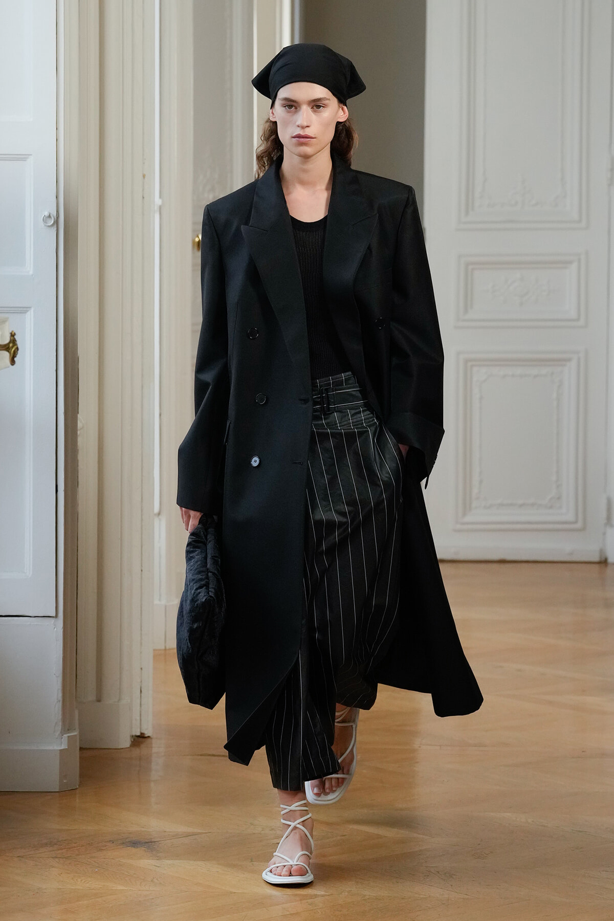 Model walking a runway in a black double‑breasted trench over a black top with pinstripe wide‑leg trousers, white strappy sandals, and a black headscarf in a white ornate room.