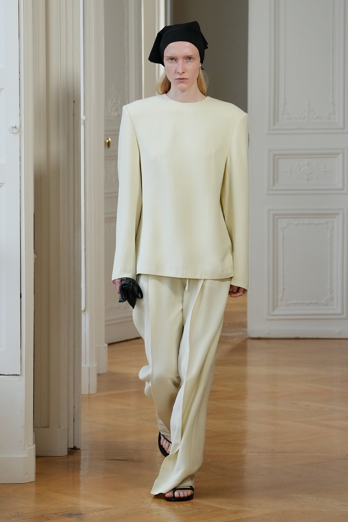 Model in a pale cream tailored suit with sculpted shoulders, black headscarf, and black sandals holding a small black clutch in a bright, ornate room user
