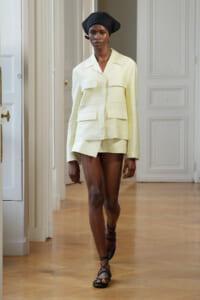 Model wearing a pale yellow utility-style jacket with large pockets and matching shorts, black headwrap, strappy sandals on a runway.