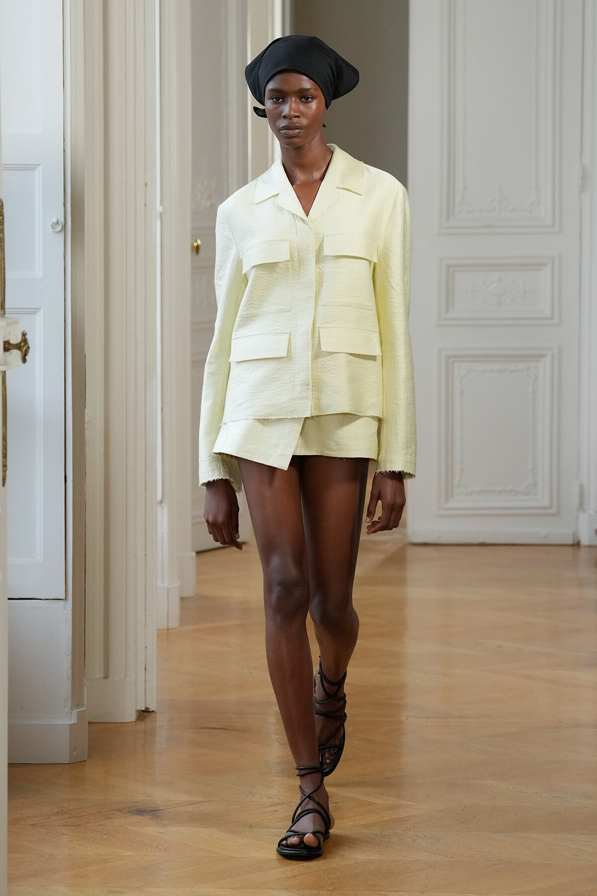 Model wearing a pale yellow utility-style jacket with large pockets and matching shorts, black headwrap, strappy sandals on a runway.