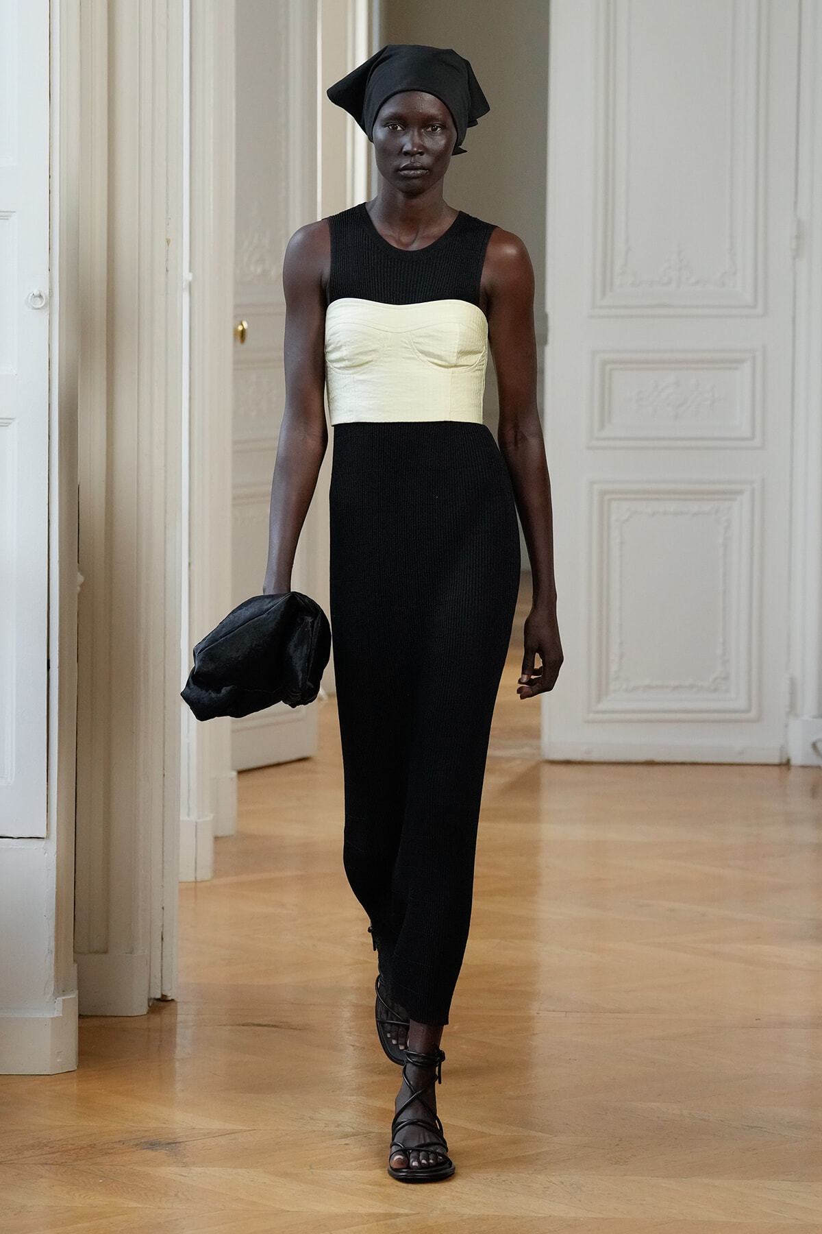 Runway model wearing a black knit dress with a cream bustier top, black headwrap, and strapped sandals, holding a black clutch in a bright room.