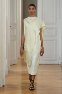 Model wearing a pale yellow draped, one-shoulder dress with a wrap skirt walking in a light-filled hallway, wearing black strappy sandals.