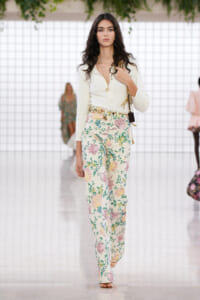 Model walking the runway in a cream blouse and floral wide-leg pants, accessorized with gold jewelry and a small shoulder bag.