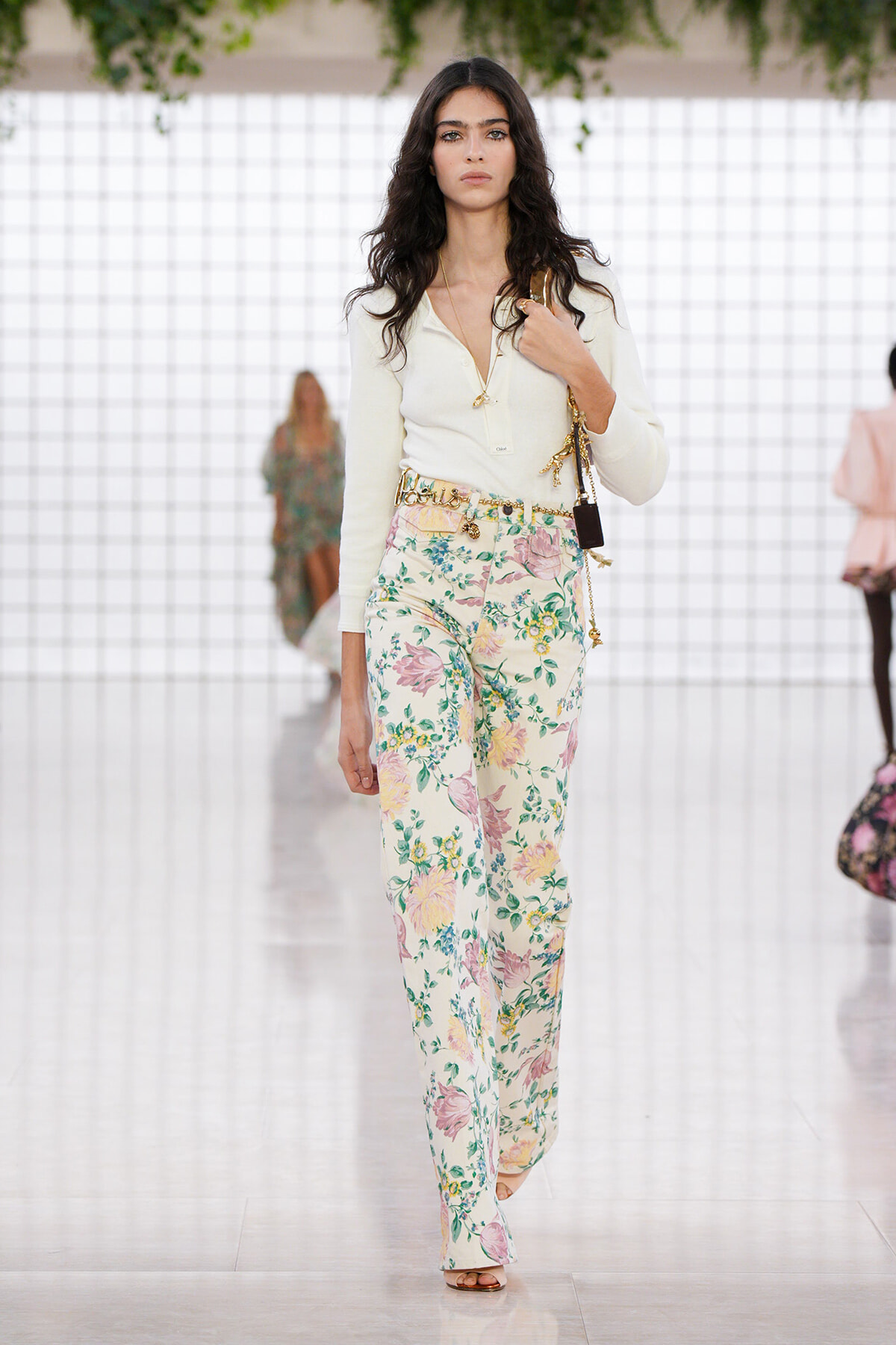 Model walking the runway in a cream blouse and floral wide-leg pants, accessorized with gold jewelry and a small shoulder bag.