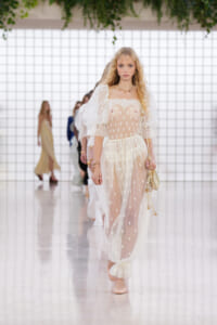 Runway model in a sheer white polka-dot gown with lace trim and puff sleeves, walking toward the camera.