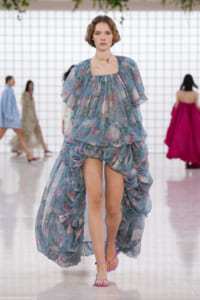 Model walking the runway in a light blue floral, voluminous gown with a dramatic high-low train and pink sandals.