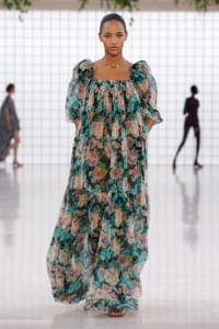 Model walking a runway in a sheer, floral maxi dress with puff sleeves in teal, pink, and cream colors.