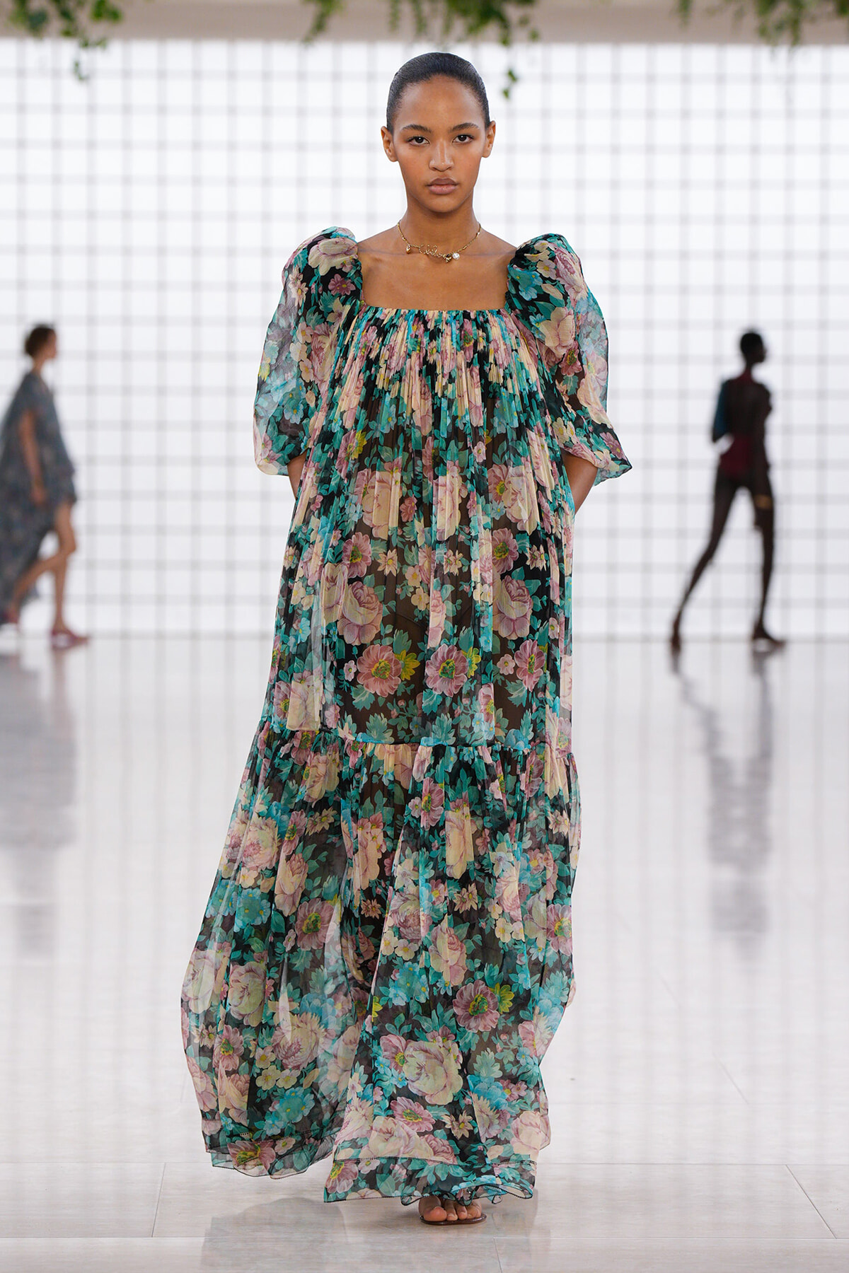 Model walking a runway in a sheer, floral maxi dress with puff sleeves in teal, pink, and cream colors.