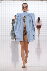 Model walks a runway in an oversized pale blue coat over a floral-knit bikini, with large sunglasses and gold earrings, strappy sandals visible in the foreground.