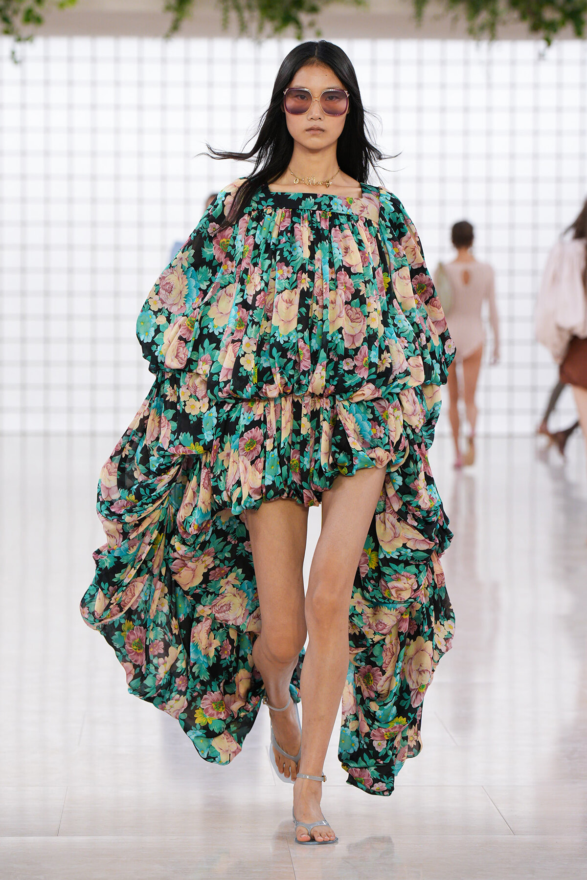 Model walking a runway in a teal floral maxi dress with pink flowers and voluminous, draped layers, wearing sunglasses.