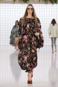 Model walking a runway in a black floral print dress with pink flowers, oversized sunglasses, and pink platform heels.
