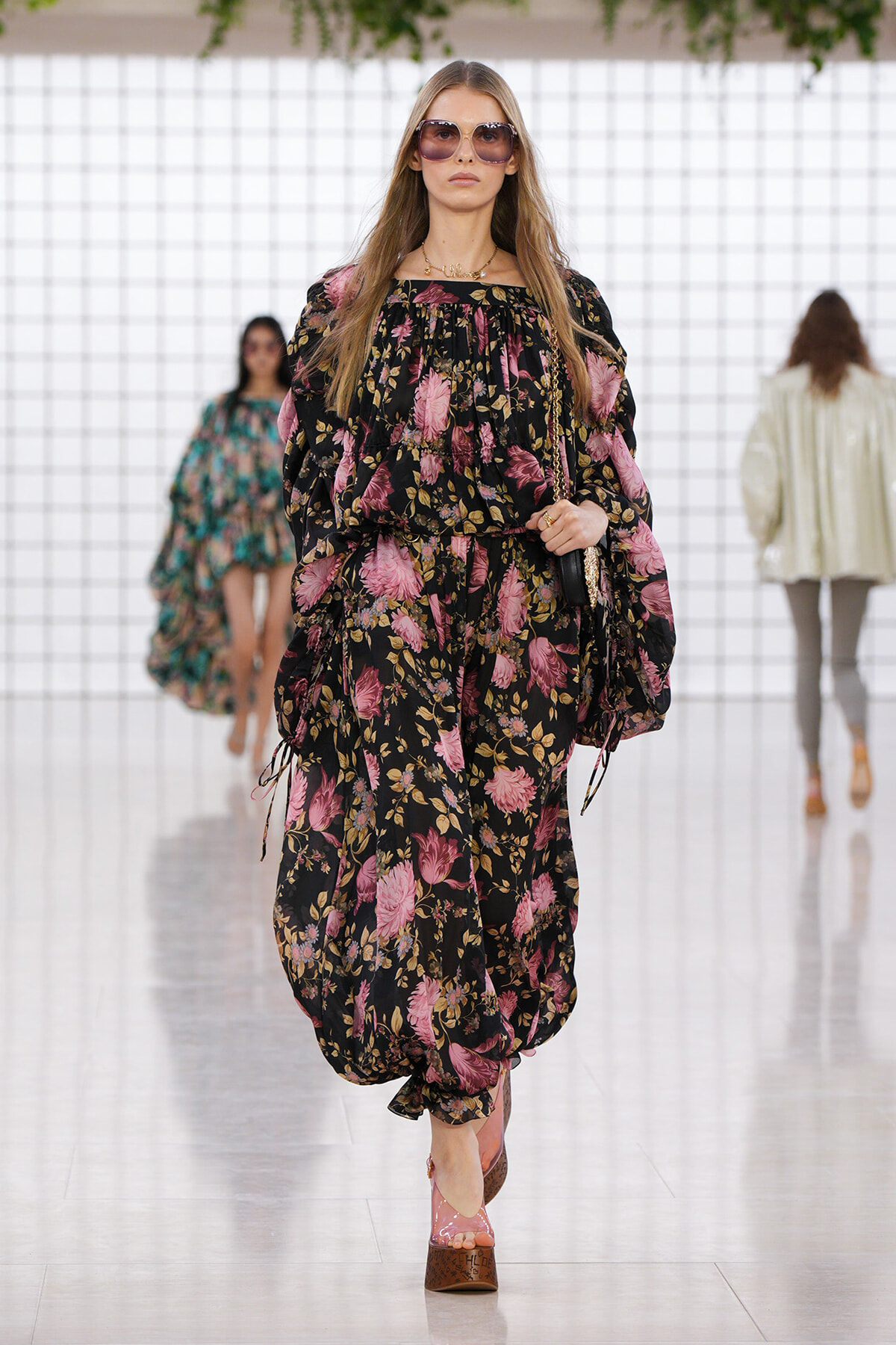 Model walking a runway in a black floral print dress with pink flowers, oversized sunglasses, and pink platform heels.