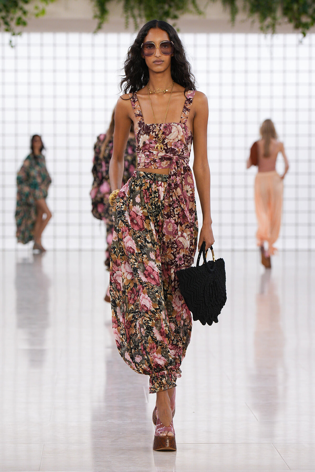 Model walking a fashion show in a pink floral two-piece outfit with a tied waist, oversized sunglasses, and a black textured handbag.