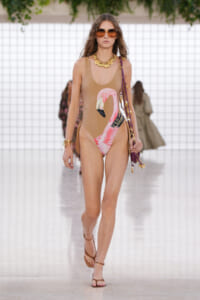 Model walking a runway in a tan one-piece swimsuit with a pink flamingo print, brown sandals, oversized sunglasses, gold jewelry, and a purple bag.