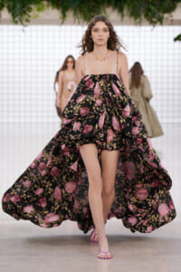 Model walking the runway in a black floral off-shoulder gown with pink flowers and billowy volume, pink sandals visible.