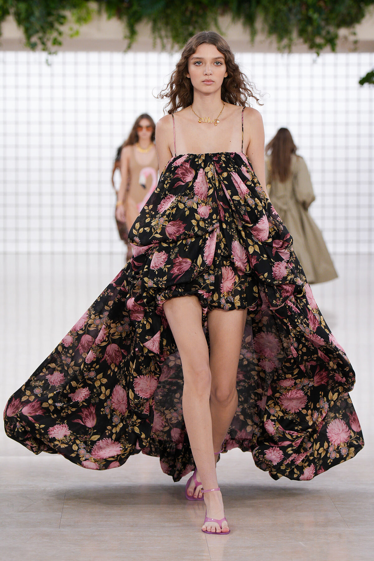 Model walking the runway in a black floral off-shoulder gown with pink flowers and billowy volume, pink sandals visible.