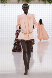 Female model wearing a peach puff-sleeve blouse over floral shorts with a gold chain belt, walking the fashion runway.