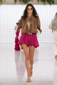 Model on runway in a brown oversized jacket over lace blouse and bright magenta bubble shorts with sunglasses on. (informative fashion image)