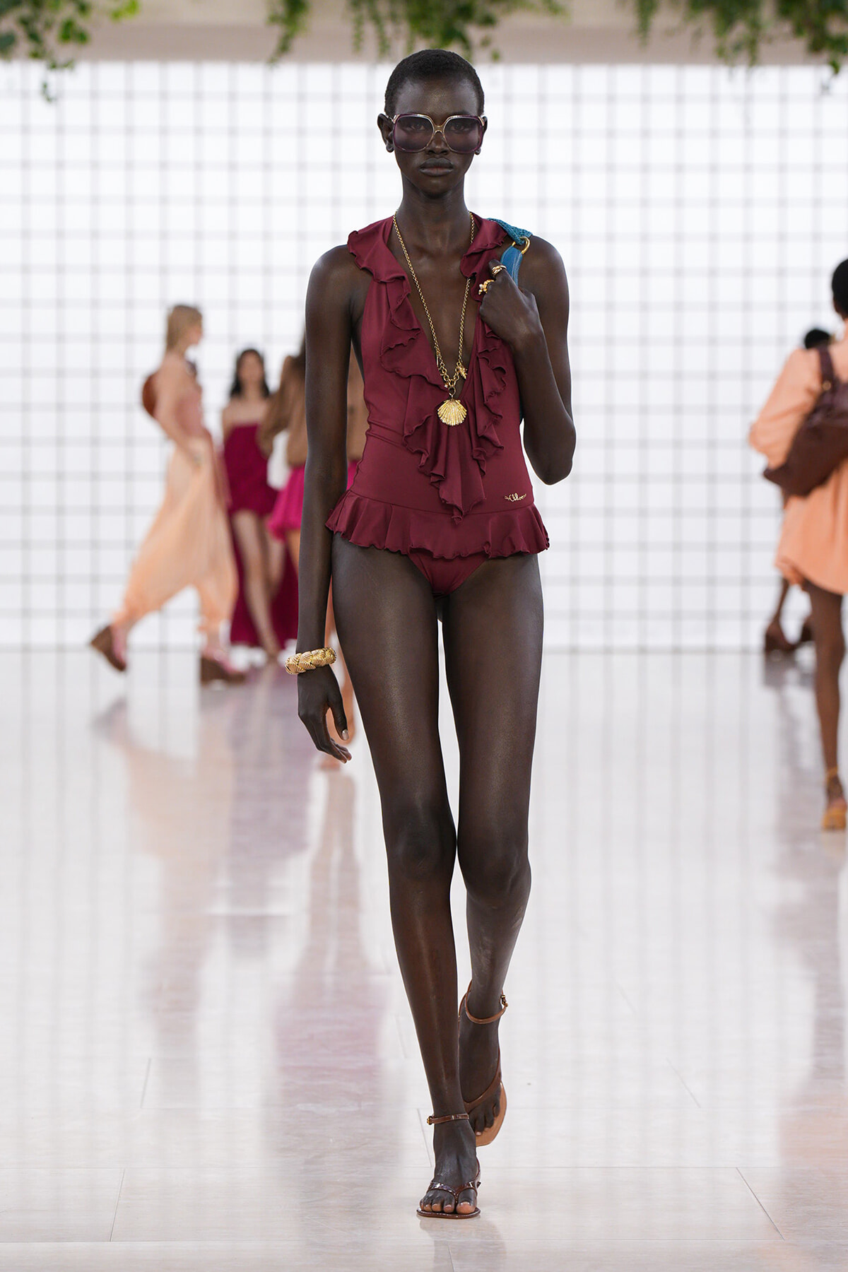 Model wearing a burgundy ruffled one-piece swimsuit with gold jewelry, sunglasses, and strappy sandals walking a fashion runway.