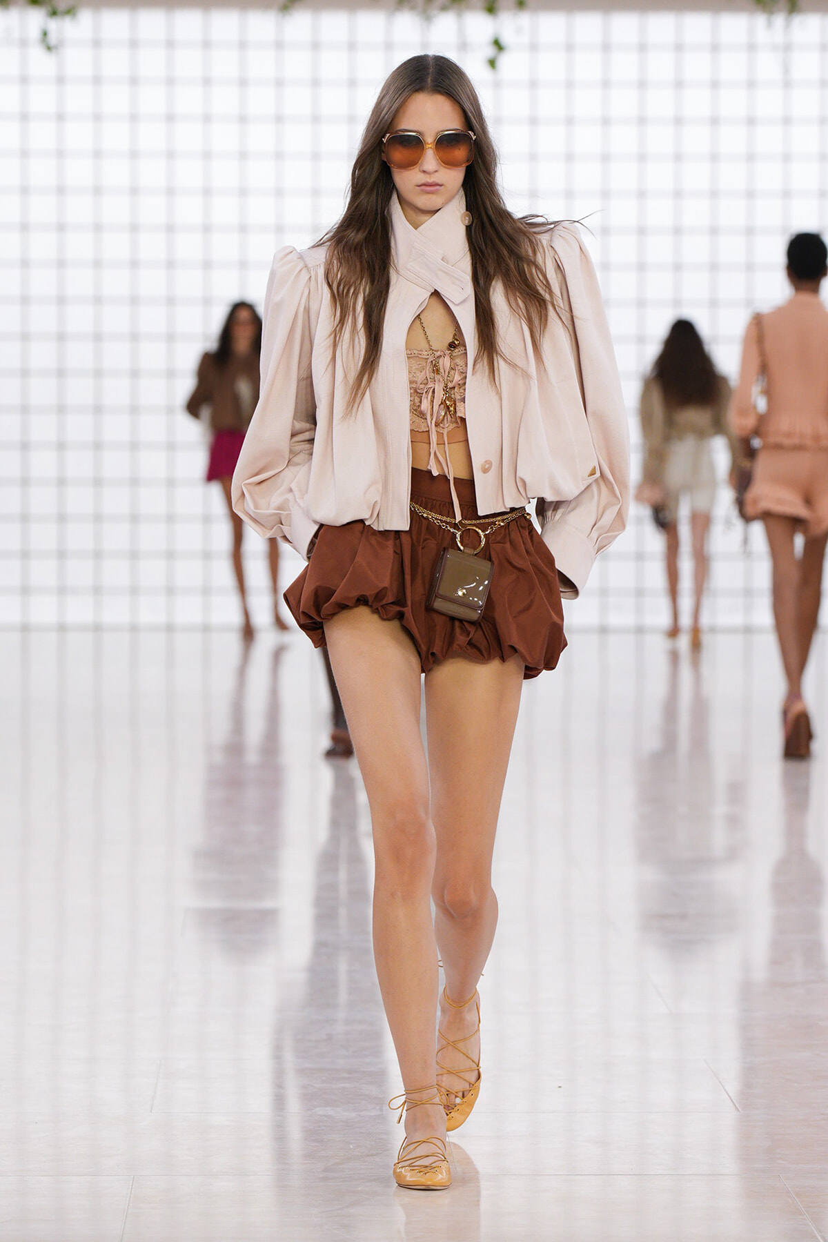 Model walks a fashion runway in a beige puff-sleeve jacket over a lace-up beige top and brown bubble shorts, with tan sandals and oversized sunglasses.