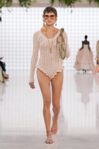 Model on a runway wearing a cream crochet bodysuit with scalloped edges, pink sandals, oversized orange-tinted sunglasses, seashell earrings, and a pale green bag.