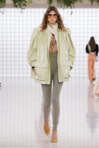 Model walking runway in oversized light-yellow glossy jacket, olive-green leggings, nude heels, and brown sunglasses