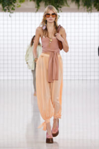 Model walks a fashion runway in a peach halter top and billowy peach pants, with oversized sunglasses and gold jewelry, carrying a brown bag.