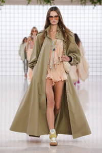 Model walking runway in an oversized olive trench over a peach lace romper, with large sunglasses and beige sneakers.