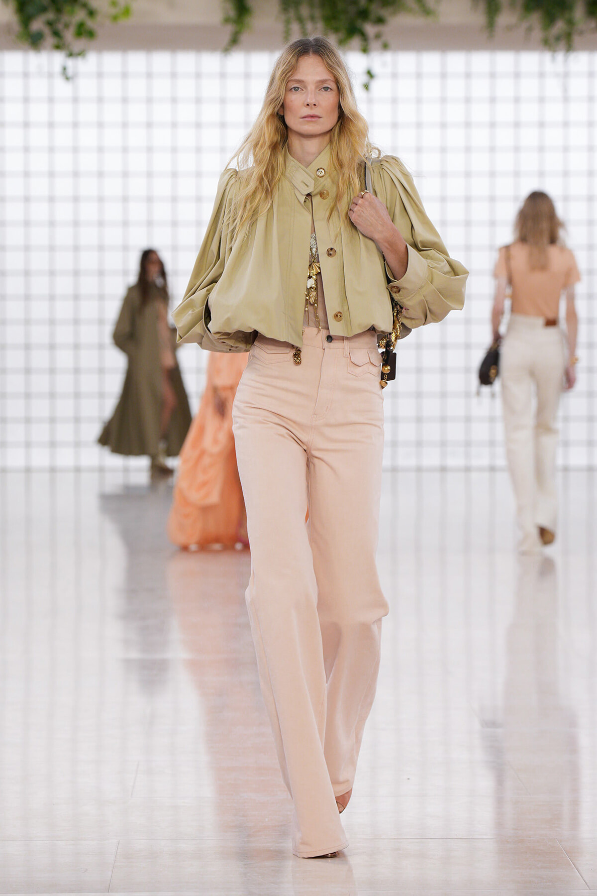 Runway model wearing a beige oversized blouse with puff sleeves and high-waisted blush wide-leg pants, walking down a glossy white catwalk.