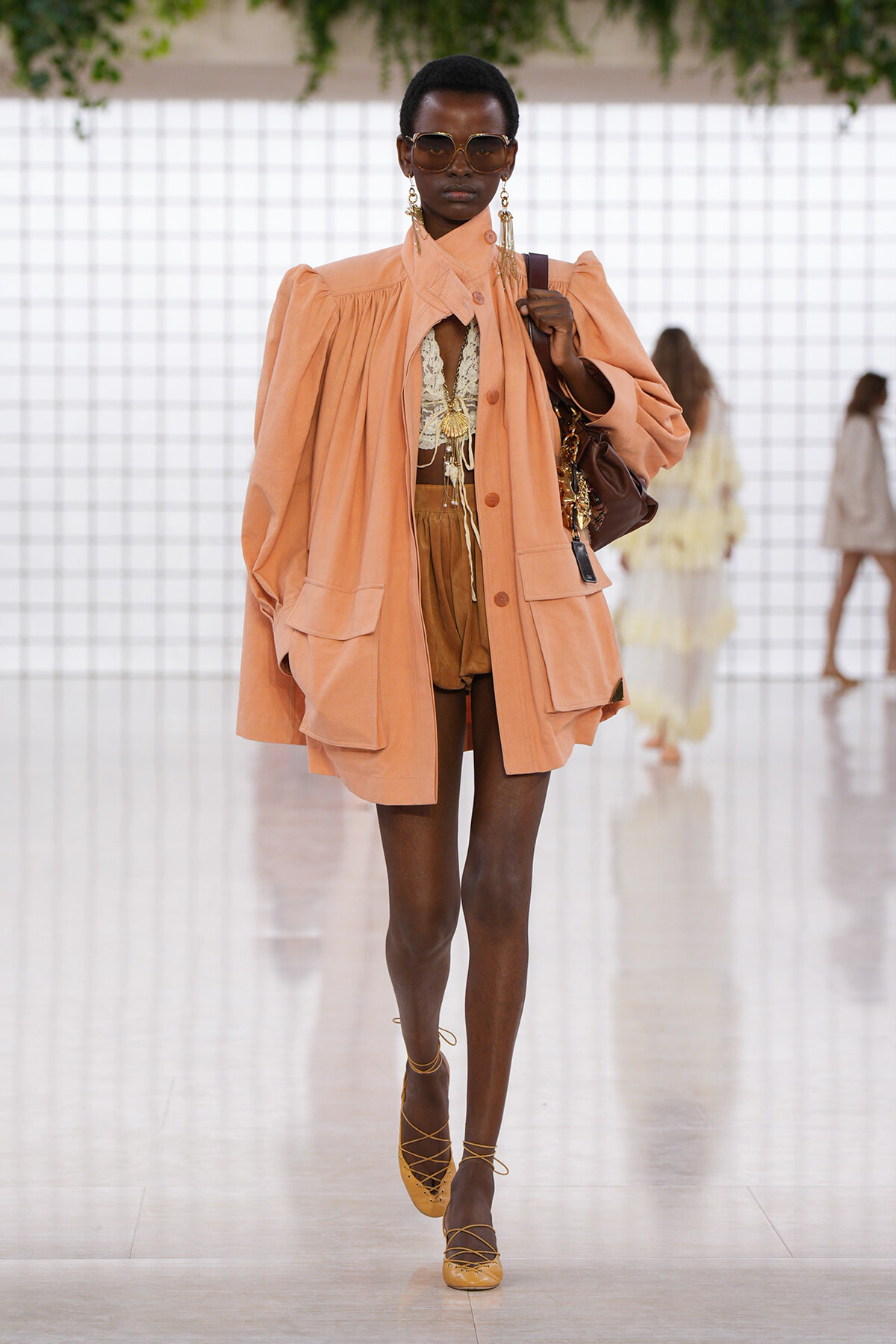 Model walks the runway in a peach oversized coat over tan shorts and a lacy top, with brown accessories and sunglasses.