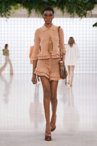 Model walks a fashion runway wearing a peach crochet two-piece set with ruffled edges, oversized sunglasses, and a brown shoulder bag.