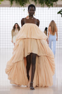 Model walking a runway in a voluminous peach tiered gown with thin straps, gold jewelry, and dark skin against a white tiled backdrop.
