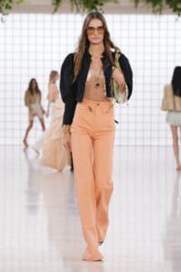 Model walking a runway in peach wide-leg pants, black cropped jacket, and lace camisole, accessorized with oversized sunglasses and gold jewelry.