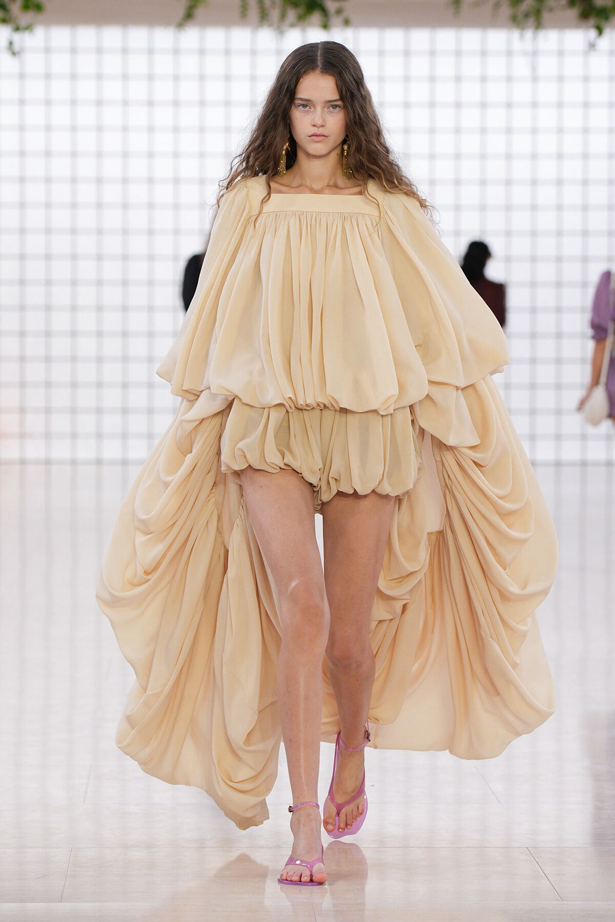Model on a runway wearing a voluminous beige off-shoulder gown with draped cape sleeves and pink sandals.