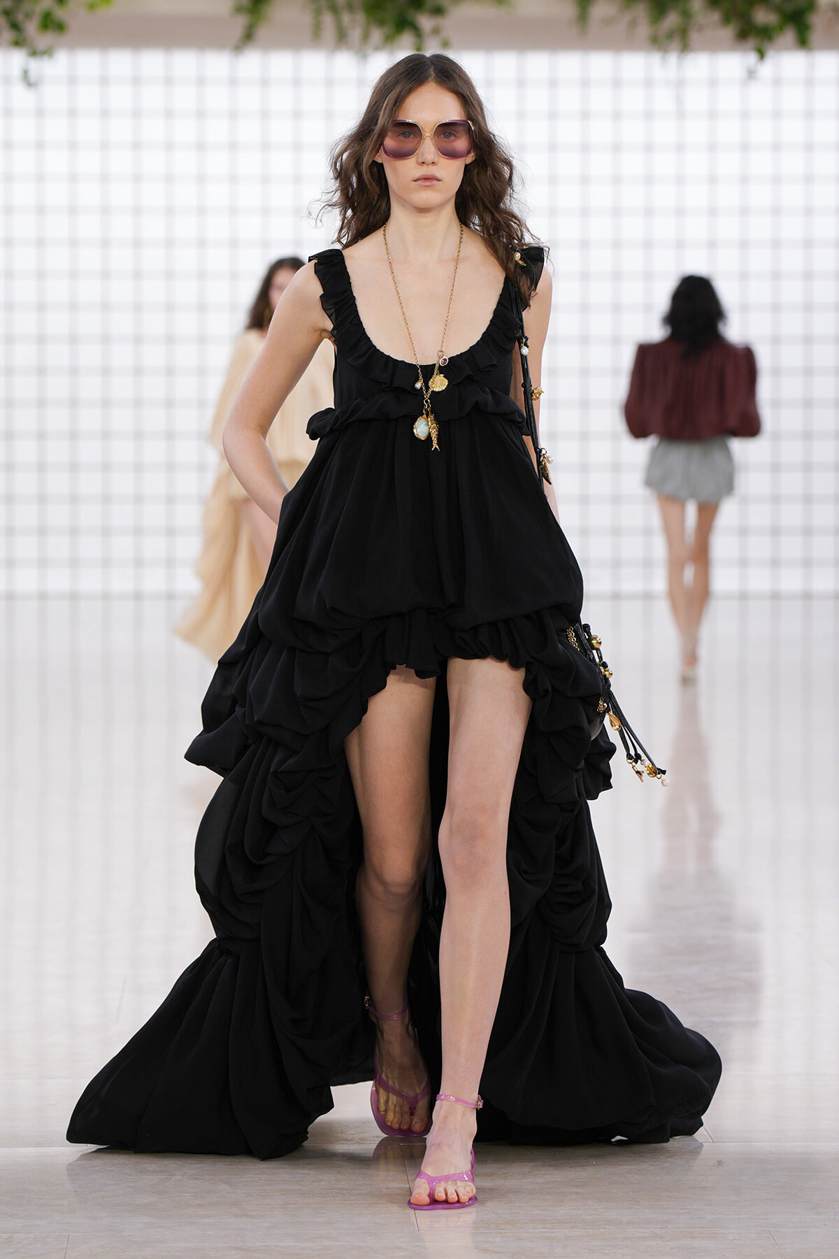 Model walks a runway in a black, layered, high-low ruffled gown with pink sunglasses and pink strappy sandals.