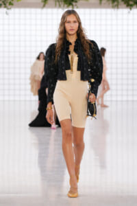 Model walking a fashion runway in a beige bodysuit with a black ruffled blouse, sheer black jacket, and tan strappy heels, carrying a small handbag.