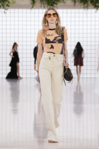 Model walking a runway in a peach graphic tee by Chloé paired with high-waisted cream wide-leg pants, oversized sunglasses, and a black crossbody bag with gold accents.