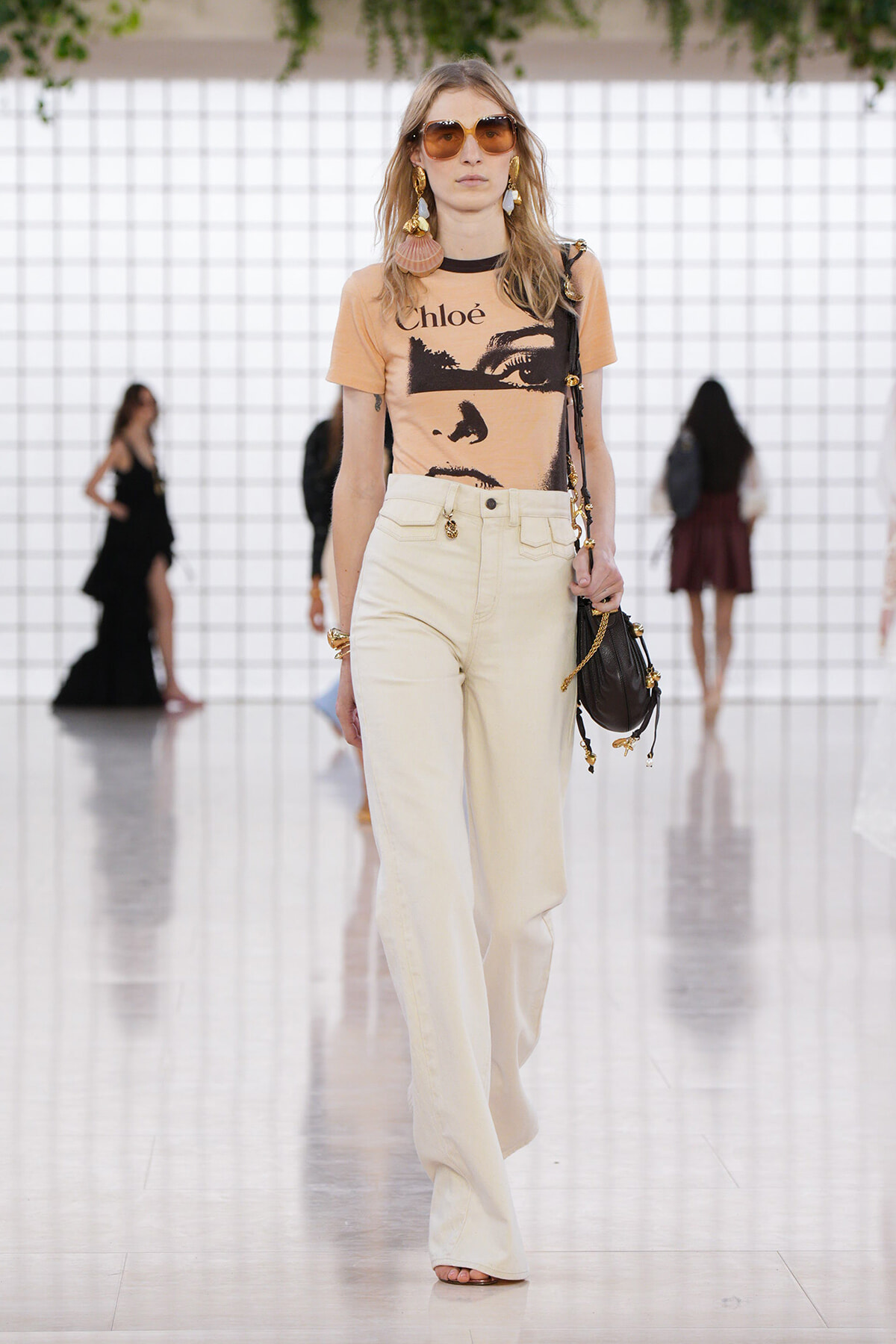 Model walking a runway in a peach graphic tee by Chloé paired with high-waisted cream wide-leg pants, oversized sunglasses, and a black crossbody bag with gold accents.