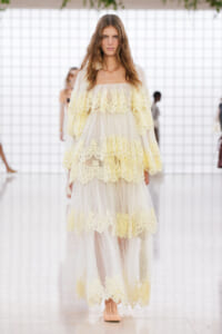 Model on a runway wearing a pale yellow lace, off-shoulder gown with sheer overlay and tiered ruffles on a translucent tulle skirt.