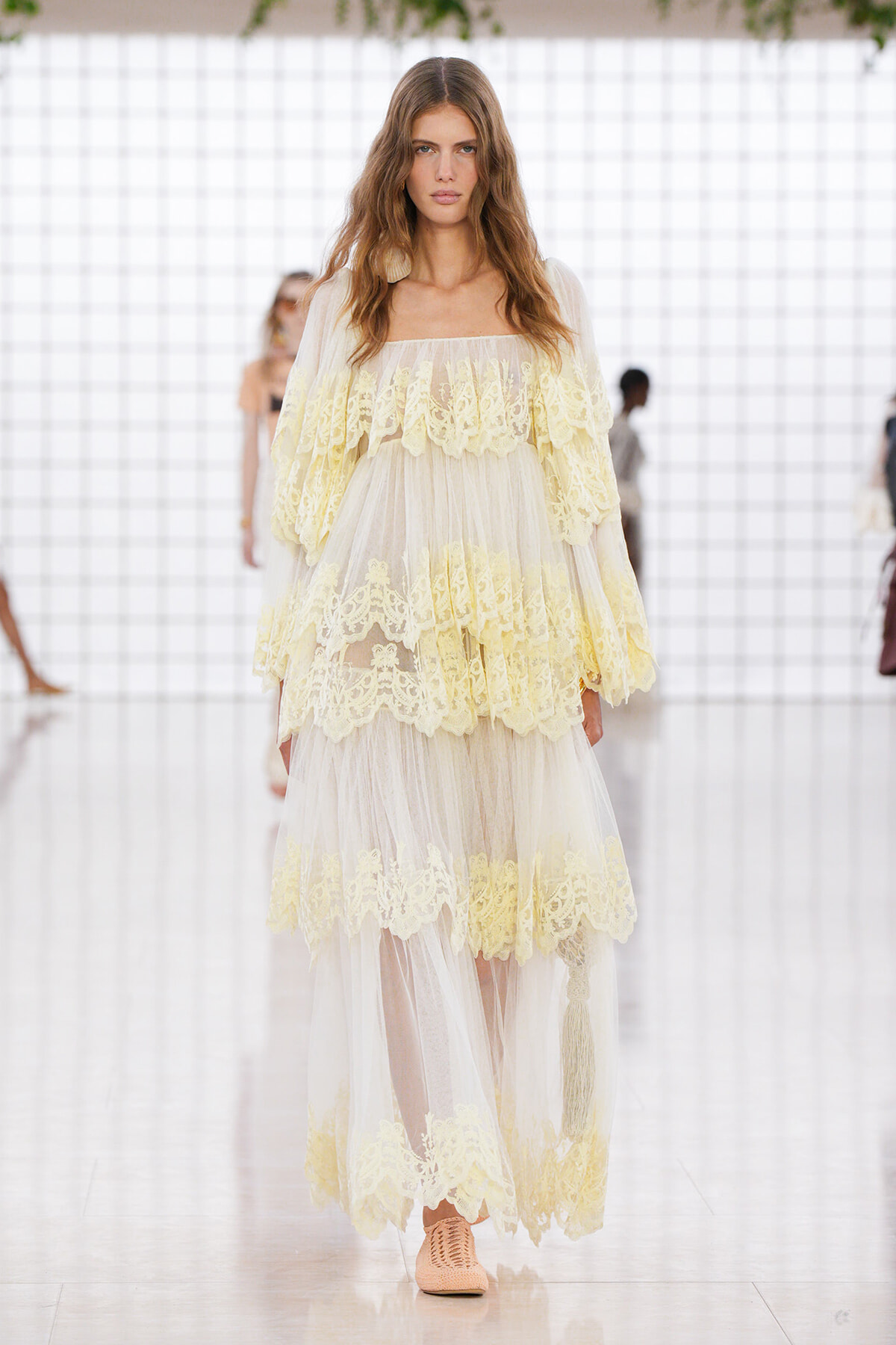 Model on a runway wearing a pale yellow lace, off-shoulder gown with sheer overlay and tiered ruffles on a translucent tulle skirt.