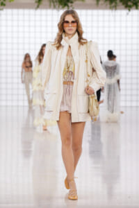 Model walking runway in an oversized cream coat over a cream lace top and pale shorts, tan sandals, and sunglasses, carrying a beige handbag.