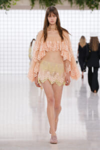 Model walking the runway in a peach lace off-shoulder cape over pale yellow lace shorts, pink flats, in a bright white venue with grid backdrop.