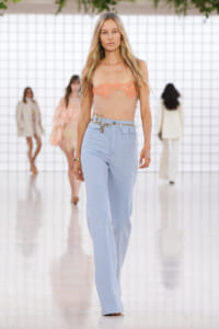 Model walks the runway in a peach lace bandeau top and light blue high-waisted wide-leg pants with a gold belt.